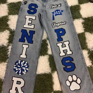 May include: Light wash denim jeans featuring custom blue and white lettering and graphics. The left leg displays the word SENIOR with a blue and white pom-pom. The right leg features Class of 2027, a graduation cap, the name Jazmine, PHS, and a paw print.