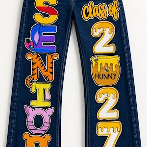 May include: Dark blue denim jeans featuring colorful character-themed lettering on the left leg spelling SENIOR and the text Class of 2027 on the right leg, with a honey pot graphic labeled HUNNY and bee illustrations integrated into the numbers.