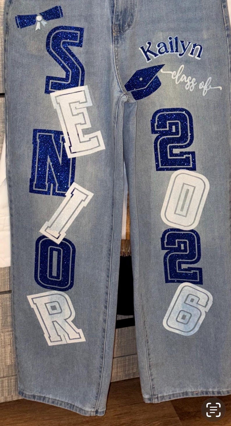 May include: Light blue denim jeans with the word "SENIOR" in blue glitter letters outlined in white. The right leg features the name "Kailyn" and "class of 2026" with a graduation cap graphic.