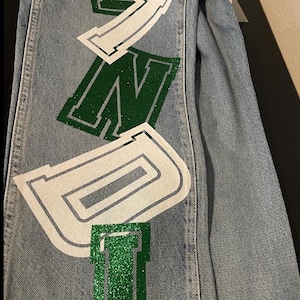 the word "BONDI" in large, green glitter letters outlined in white. The jeans are folded, showing the design on the leg. The jeans have a classic five-pocket design.
