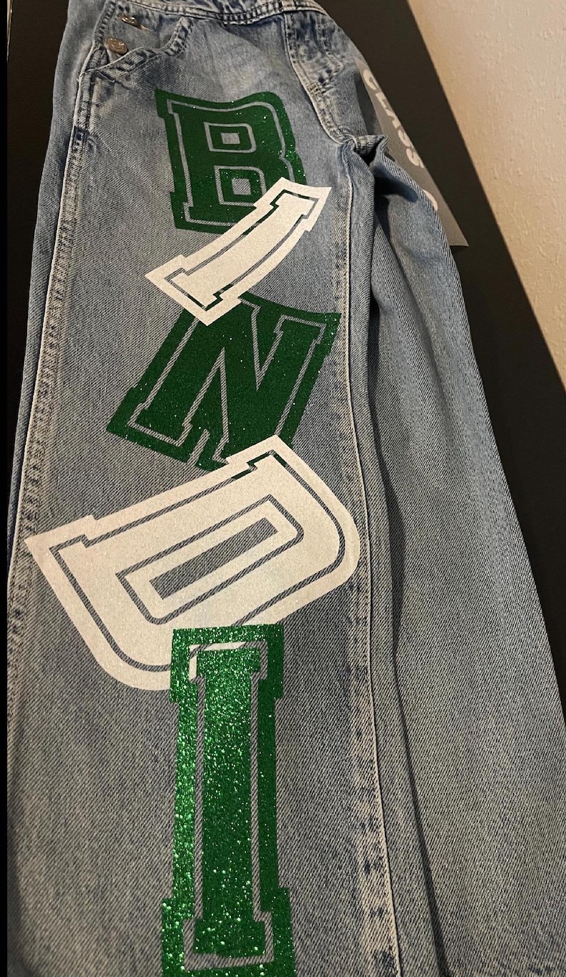 the word "BONDI" in large, green glitter letters outlined in white. The jeans are folded, showing the design on the leg. The jeans have a classic five-pocket design.
