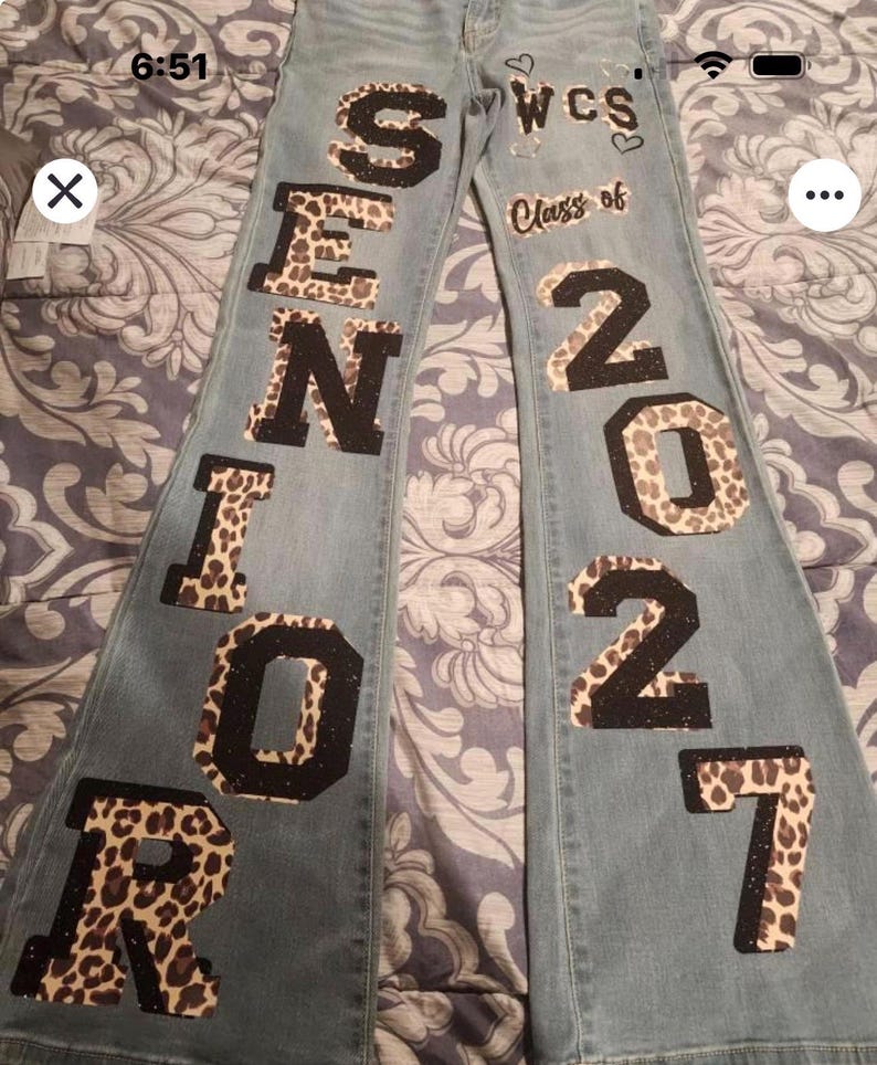 May include: Light blue denim jeans featuring large, block-style lettering down the legs. The left leg displays the word SENIOR, while the right leg shows the text Class of 2027. The letters and numbers have a black and leopard print pattern.