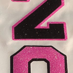 May include: A graphic design featuring the number 20. The number 2 is black with a bright pink glitter outline, and the number 0 is bright pink glitter with a black outline. The design is set against a plain, light-colored background surface.