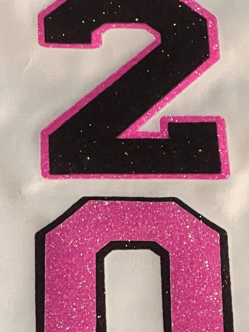 May include: A graphic design featuring the number 20. The number 2 is black with a bright pink glitter outline, and the number 0 is bright pink glitter with a black outline. The design is set against a plain, light-colored background surface.