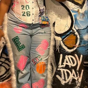 May include: A white corset top with pink accents and the numbers "20 26". Light blue jeans with pink and green painted designs, including letters, numbers, and a basketball. The outfit is trimmed with white faux fur.