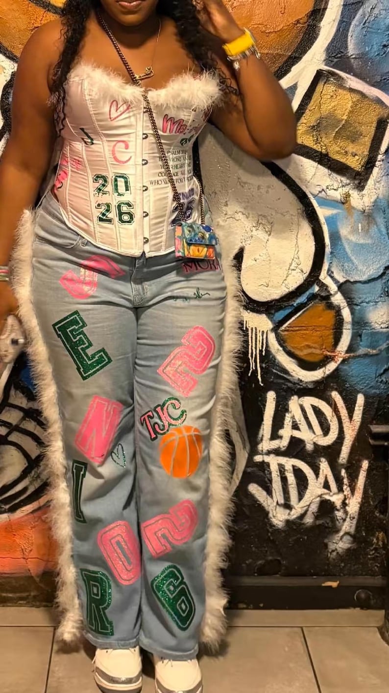 May include: A white corset top with pink accents and the numbers "20 26". Light blue jeans with pink and green painted designs, including letters, numbers, and a basketball. The outfit is trimmed with white faux fur.