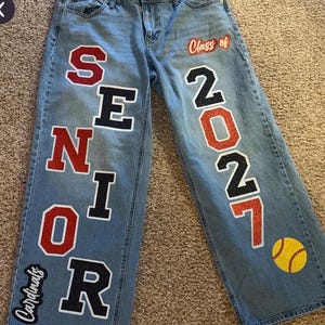 May include: Light blue denim jeans featuring large, glittery varsity-style lettering that spells SENIOR down the left leg and the numbers 2027 down the right leg. The text Class of is above the numbers, and a yellow softball and Cardinals logo appear.