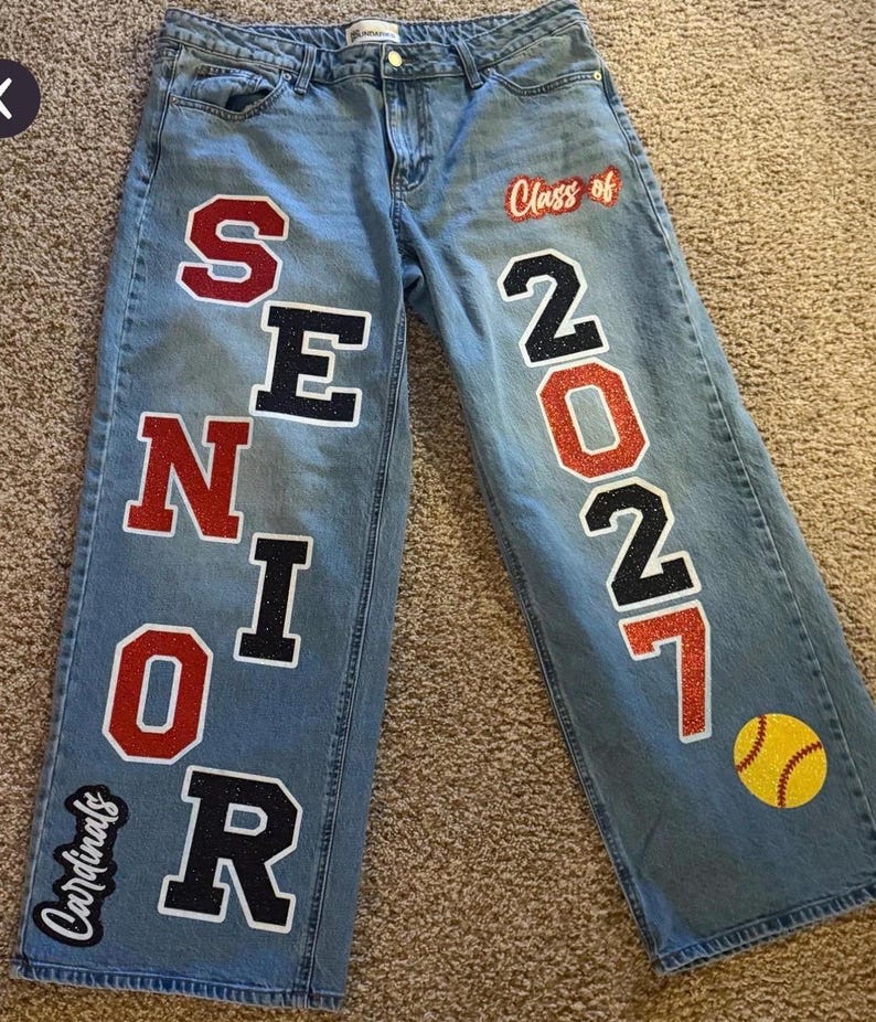 May include: Light blue denim jeans featuring large, glittery varsity-style lettering that spells SENIOR down the left leg and the numbers 2027 down the right leg. The text Class of is above the numbers, and a yellow softball and Cardinals logo appear.