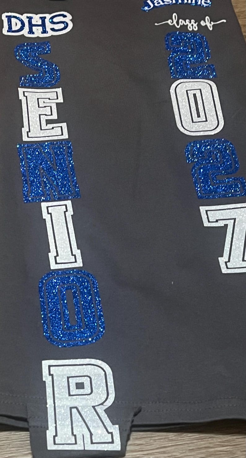 May include: Gray fabric with the text "DHS SENIOR 2023" in white and blue glitter lettering. The name "Jasmine" and "class of" are also visible in the image.