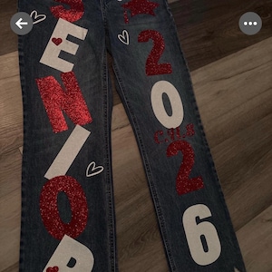 May include: Blue denim jeans with distressed hems, featuring the word "SENIOR" in white and red glitter lettering. The jeans also display "CLASS OF 2026" with a graduation cap graphic and hearts.