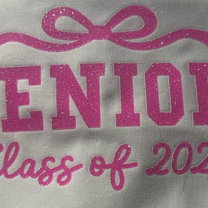 May include: White fabric with the words "SENIOR Class of 2026" in pink glitter lettering. A pink glitter bow is above the word "SENIOR".