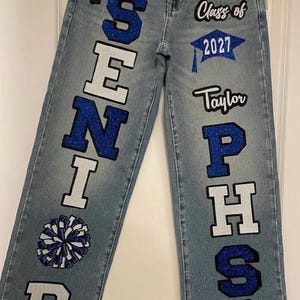 May include: Light blue denim jeans featuring custom blue glitter lettering and graphics. The left leg displays the word SENIOR with a paw print and a pom-pom. The right leg features Class of 2027, a graduation cap, the name Taylor, PHS, and a paw print.