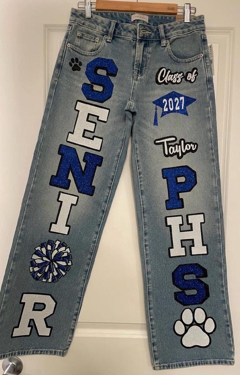 May include: Light blue denim jeans featuring custom blue glitter lettering and graphics. The left leg displays the word SENIOR with a paw print and a pom-pom. The right leg features Class of 2027, a graduation cap, the name Taylor, PHS, and a paw print.
