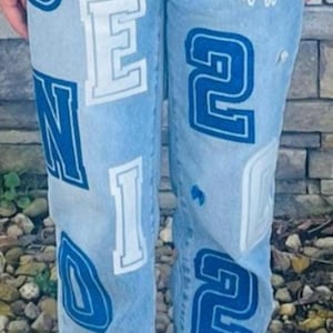 May include: Light blue denim jeans with white and blue lettering and graphics. The jeans feature the words "SENIOR", "2022", and "Class of" along with the school name "Chrystian SHS".