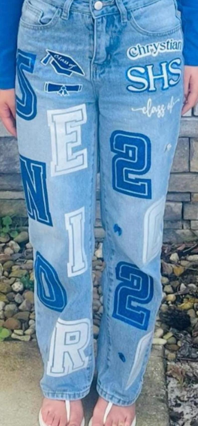 May include: Light blue denim jeans with white and blue lettering and graphics. The jeans feature the words "SENIOR", "2022", and "Class of" along with the school name "Chrystian SHS".