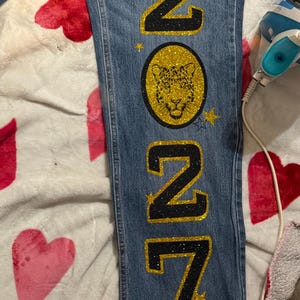 May include: Blue denim jeans featuring a vertical design with the glittery black and gold text 2027. A circular gold glitter patch displays a black leopard face graphic. Small gold and blue glitter stars are positioned around the text and patch.