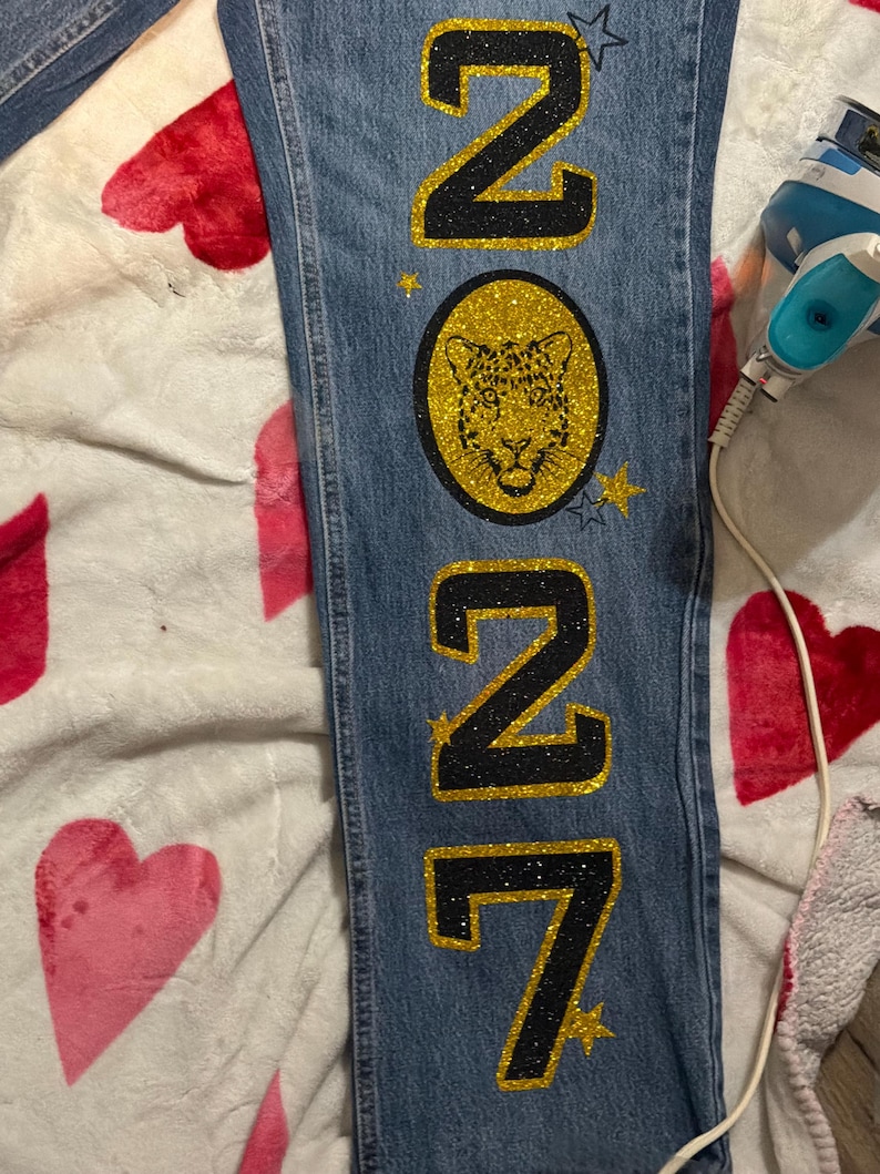 May include: Blue denim jeans featuring a vertical design with the glittery black and gold text 2027. A circular gold glitter patch displays a black leopard face graphic. Small gold and blue glitter stars are positioned around the text and patch.