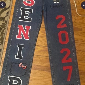 May include: Dark blue denim jeans featuring the word SENIOR in red and black block letters, the text Class of 2027, and the number 2027 in red. The jeans include a Hello Kitty graphic, small silver studs along the seams, and the text GOD DID.