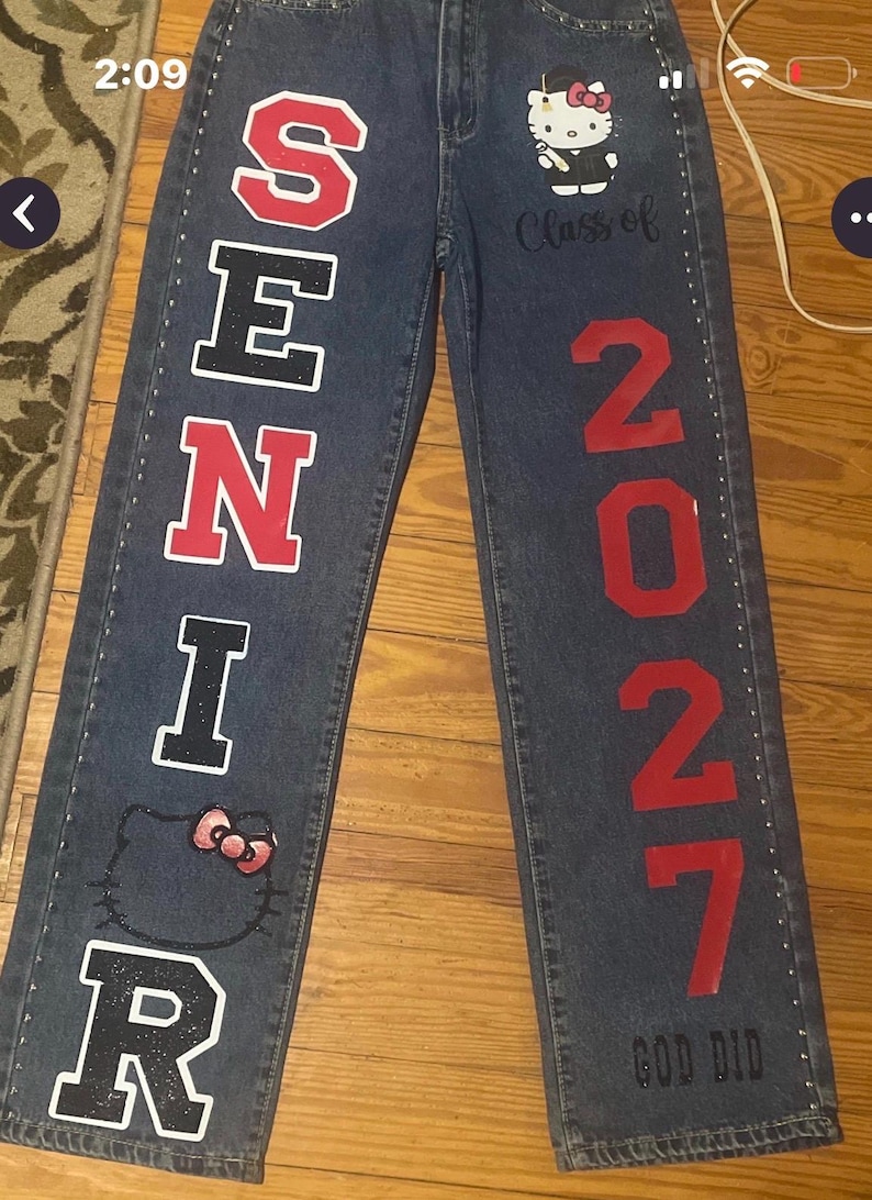 May include: Dark blue denim jeans featuring the word SENIOR in red and black block letters, the text Class of 2027, and the number 2027 in red. The jeans include a Hello Kitty graphic, small silver studs along the seams, and the text GOD DID.