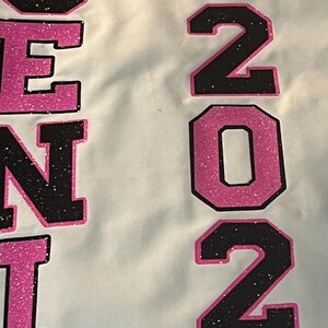 May include: A white fabric surface featuring the text "SENIOR" and "2024" in large, block-style lettering. The letters and numbers are filled with a black glitter texture and outlined in bright pink glitter. A pink glitter graduation cap icon sits above the text "CLASS OF".