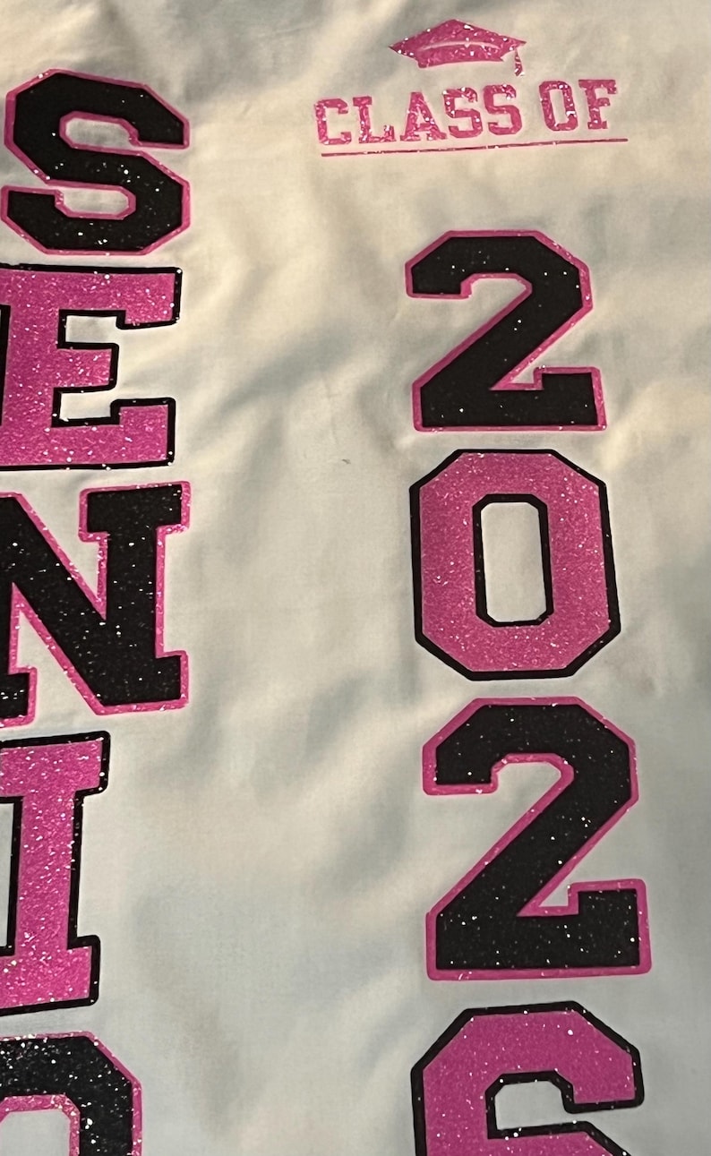 May include: A white fabric surface featuring the text "SENIOR" and "2024" in large, block-style lettering. The letters and numbers are filled with a black glitter texture and outlined in bright pink glitter. A pink glitter graduation cap icon sits above the text "CLASS OF".