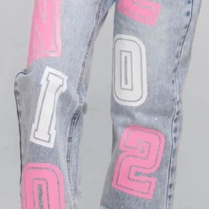 May include: Light wash denim jeans with pink and white lettering spelling "SENIOR 2026" on the legs. The words "class of" are written in cursive above the right pocket. Rhinestones are scattered throughout the jeans.