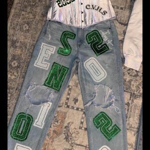 May include: A silver corset top and light wash denim jeans with distressed details. The jeans feature green and white lettering and numbers, including "2026" and "CVHS". A graduation cap graphic is also visible.