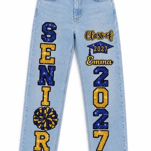 May include: Light blue denim jeans featuring vertical text and graphics. The left leg displays the word SENIOR in blue and gold glitter with a pom-pom graphic. The right leg features Class of 2027, Emma, 2027, and Riders in blue and gold glitter.