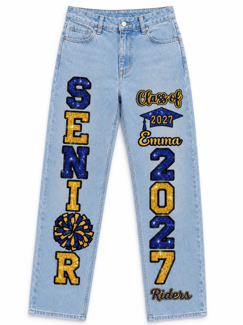 May include: Light blue denim jeans featuring vertical text and graphics. The left leg displays the word SENIOR in blue and gold glitter with a pom-pom graphic. The right leg features Class of 2027, Emma, 2027, and Riders in blue and gold glitter.