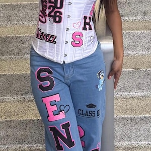 May include: A white strapless corset top featuring pink and black text that reads Senior, Class of 26, and Kenzz, paired with light blue denim jeans featuring the word Senior in large pink and black letters, a graduation cap icon, and a cartoon character.