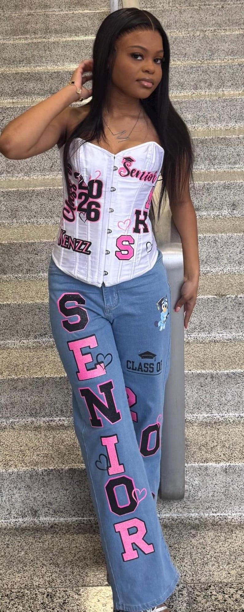 May include: A white strapless corset top featuring pink and black text that reads Senior, Class of 26, and Kenzz, paired with light blue denim jeans featuring the word Senior in large pink and black letters, a graduation cap icon, and a cartoon character.
