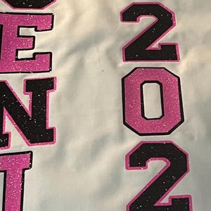May include: A white fabric surface featuring the text "SENIOR" and "2024" in large, block-style lettering. The letters and numbers are filled with a black glitter texture and outlined in bright pink glitter. A pink glitter graduation cap icon sits above the text "CLASS OF".