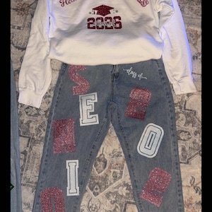 May include: A white sweatshirt and denim jeans set. The sweatshirt has the word "Heaven" and "CVHS" printed on it, along with a graduation cap and the year "2026". The jeans have the word "SENIOR" and the number "26" on them.