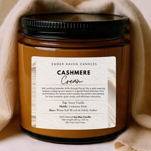以下が含まれることがあります： 黒い蓋が付いた茶色のガラス瓶のキャンドルで、「Cashmere Cream」と表示されています。ラベルには、バニラ、カシミアムスク、アンバーの香りのノートが記載されています。ベージュの生地がキャンドルの周りにドレープされています。