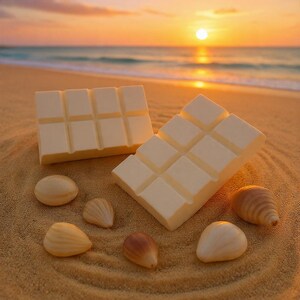 Peut inclure: Deux barres de chocolat blanc avec des segments carrés sont disposées sur une plage de sable, à côté de plusieurs coquillages. La scène est baignée par la lumière chaude d'un coucher de soleil sur l'océan.