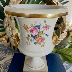 May include: A white ceramic vase with a wide rim and gold trim. The vase features a floral design with pink, blue, and yellow flowers. It has gold handles and sits on a square base.