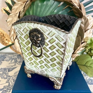 May include: A decorative planter with a distressed green and white woven pattern. It features a bronze lion's head with a ring handle and ornate feet. The interior is black.