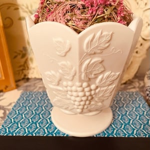 May include: A white, vintage-style vase with a grape and leaf design, filled with dried pink flowers and grasses. The vase has a scalloped top and a pedestal base, and is sitting on a blue patterned surface.