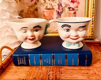 Könnte beinhalten: Zwei Vintage-Sahnekännchen von Baileys aus Keramik mit lächelnden Gesichtern, eines mit Henkel und das andere mit Fliege, stehen auf einem blauen Buch. Die Sahnekännchen haben das Baileys-Logo und stehen auf einem Bambustablett.