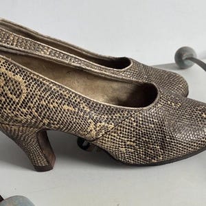 May include: A pair of slip-on court shoes featuring a textured, snakeskin-patterned exterior in shades of brown, tan, and cream. These shoes have a rounded toe, a low-profile heel, and a neutral-toned interior lining, showcasing a classic silhouette.