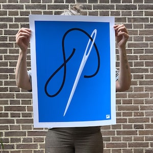 May include: A blue poster with a white border features a minimalist design of a needle and thread. The needle is white, and the thread is black, creating a striking contrast against the blue background. The poster is held up in front of a brick wall.