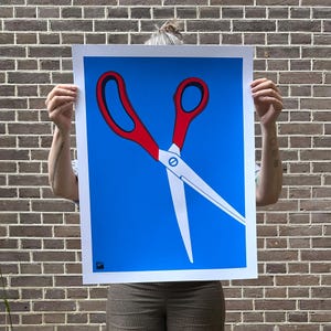 May include: A large poster featuring a stylized illustration of red scissors against a bright blue background. The scissors are depicted in a bold, graphic style, with white blades and a red handle. The poster is held up in front of a brick wall.