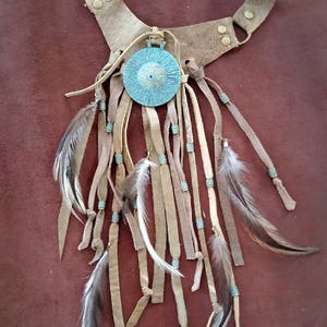 Puede incluir: Un collar de cuero marrón con un colgante de metal turquesa y flecos. Los flecos están hechos de cuero y plumas.