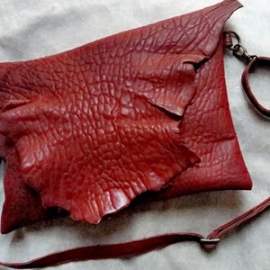 Pu&ograve; includere: Una borsa a tracolla in pelle rossa con una superficie testurizzata e granulosa. La borsa ha una chiusura a lembo e una lunga tracolla regolabile.