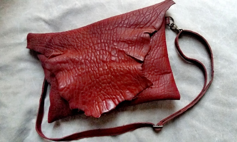 Pu&ograve; includere: Una borsa a tracolla in pelle rossa con una superficie testurizzata e granulosa. La borsa ha una chiusura a lembo e una lunga tracolla regolabile.
