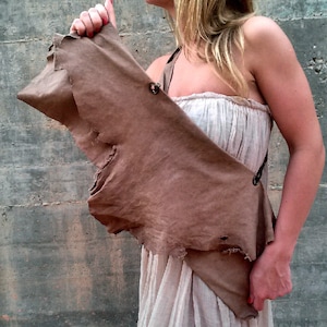 Puede incluir: Un bolso de cuero marrón con un borde deshilachado y una correa. El bolso se lleva sobre el hombro de una persona que lleva un vestido blanco.