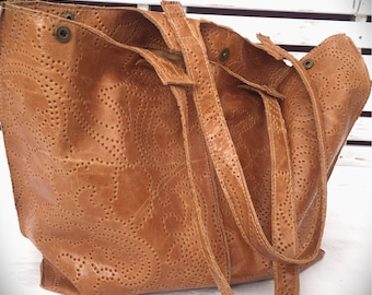 May include: A brown leather tote bag with a unique patterned design. The bag has two long straps and a top zipper closure.