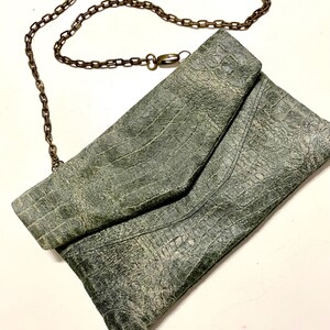 Puede incluir: Un bolso de mano de cuero verde con una correa de cadena. El bolso tiene un diseño texturizado, con estampado de cocodrilo.