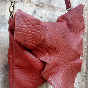 Pu&ograve; includere: Una borsa a tracolla in pelle rossa con una superficie strutturata e una chiusura a lembo. La borsa ha una tracolla lunga e una chiusura in metallo.