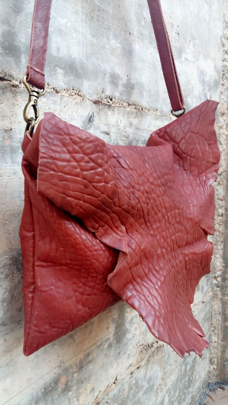Pu&ograve; includere: Una borsa a tracolla in pelle rossa con una superficie strutturata e una chiusura a lembo. La borsa ha una tracolla lunga e una chiusura in metallo.